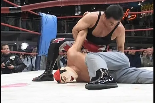 Video still of a wrestling match with a masked man pinned to the ground 