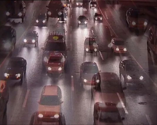 A video still depicting a traffic filled road, each car with headlights illuminating the night
