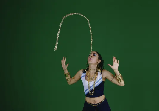 Against a green background, a woman stands with hands up, spitting a gold chain into the air like a fountain of water