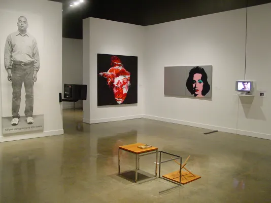 Gallery space including 2 video monitors and 3 large works mounted on the walls and small end tables centered in the room