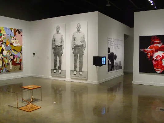 Multiple large works and a box monitor hang on the walls. In the center of the gallery, there are 2 small tables, 1 tipped over