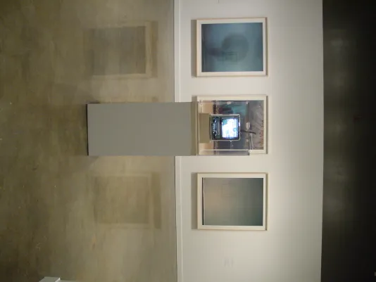 3 framed images hang behind a vitrine housing a miniature television with a glowing screen.