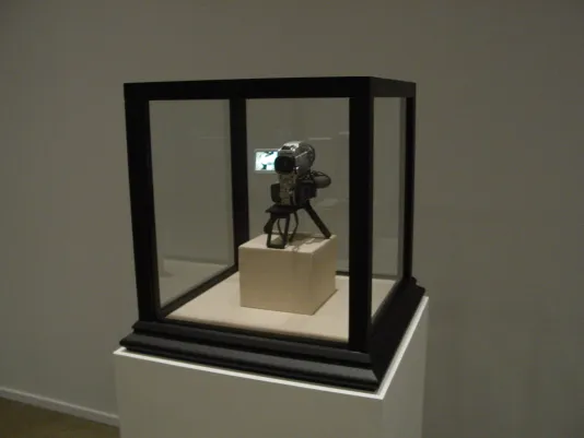 A digital camcorder sits on a pedestal within a vitrine, framed with black edges.