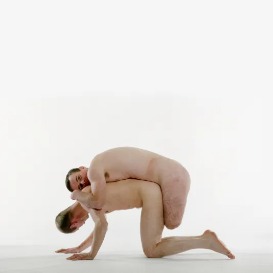 2 naked men, one on his hands and knees and the other, without legs, hangs on his back looking towards the camera