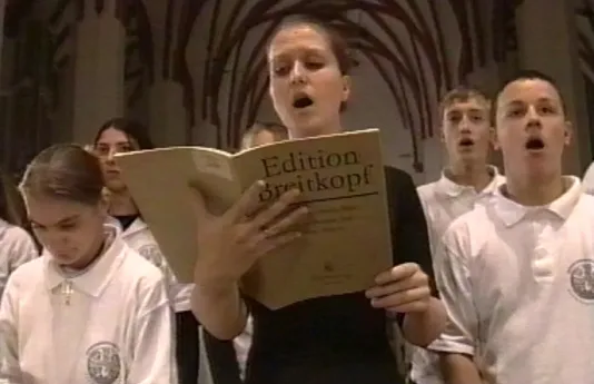 A singing choir of young people wearing white polo shirts. One woman, wearing black, stands in the center holding a song book
