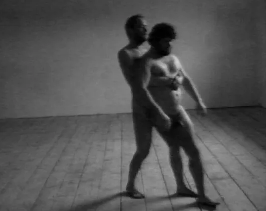 Black and white still of 2 unclothed men in an empty room. One without his right leg, is being held up by the man behind him