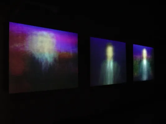 Three videos projected in a dark room show blurred images of men in suits in the middle of each frame.