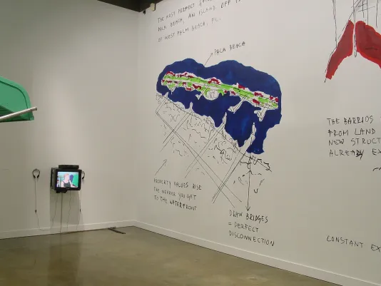 In the corner of a white gallery space, a small box monitor is mounted to the right of large scale wall drawings and writings