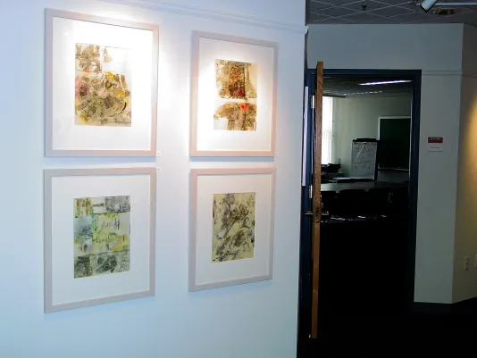 Four framed abstract artworks are hung in a grid on a wall outside of a conference room with a long table and chairs.