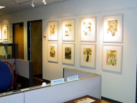 Eight framed abstract artworks are hung in a grid on a wall right across from a cubicle in the Dean's gallery office space.