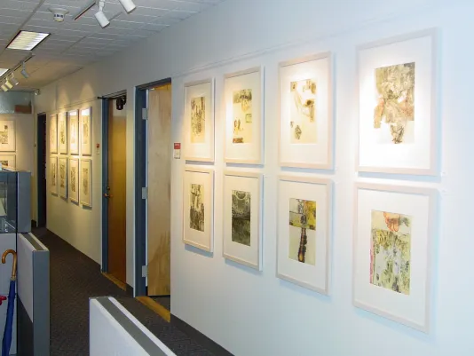 A view of a hallway in an office space where many brightly lit framed abstract artworks hang in grids across from cubicles.