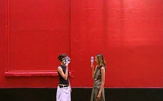 Video still from Runa Islam of two women, facing each other, hiding behind paper face masks in front of a red wall