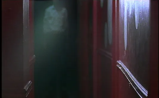 Video still from Runa Islam shows a dark red hallway leading to a blurry male figure in the background