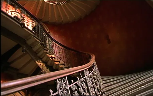 Video still shot from the bottom of an ornate winding staircase looks up at rust colored patterned wallpaper