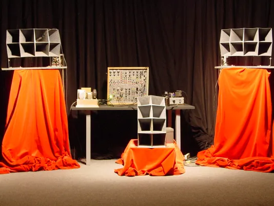 In front of black curtains, a movie-palace sound system is spread across a table and 3 pedestals draped with orange fabric