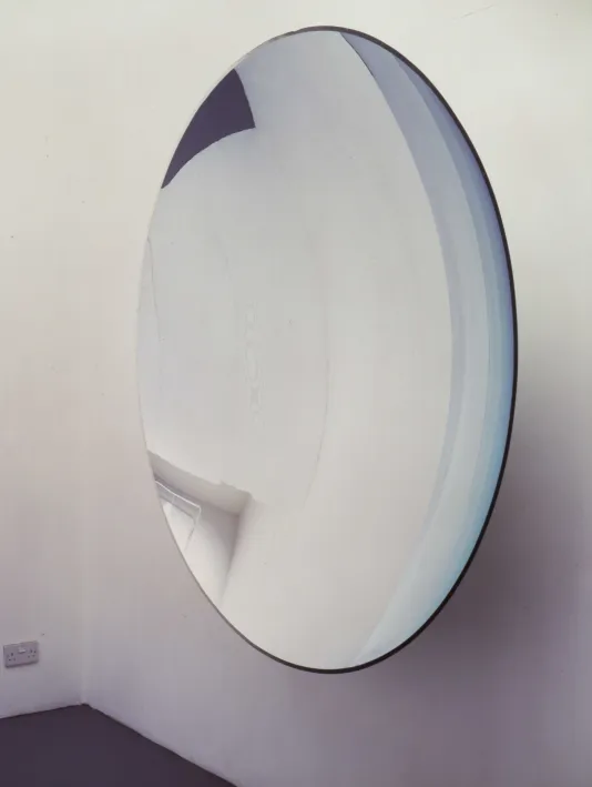 Close view of a large concave mirrored sculpture hung on a wall and reflecting the surrounding gallery space.