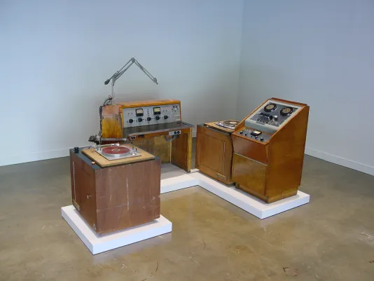 Vintage wood paneled radio broadcast equipment installed on low white pedestals on the floor in the corner of the gallery.