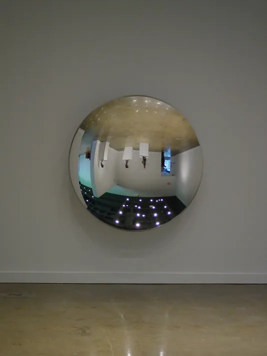 Direct view of a large circular shaped mirror sculpture hung on a wall reflecting its surrounding gallery space upside down.
