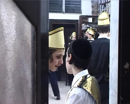 A young boy wearing a yarmulka watches from a doorway as carousing men dance in the street as part of the feast of Purim.