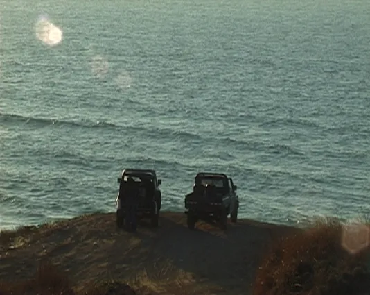 Two off road vehicles are parked at the edge of a cliff overlooking an ocean, a person stands at the rear of one of them.