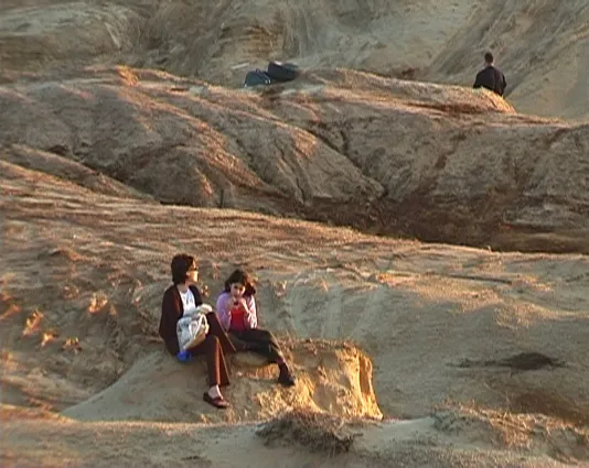 A woman and child sit on a sandy coastal hill outside of Tel Aviv, near an area where men drive trucks off road. 