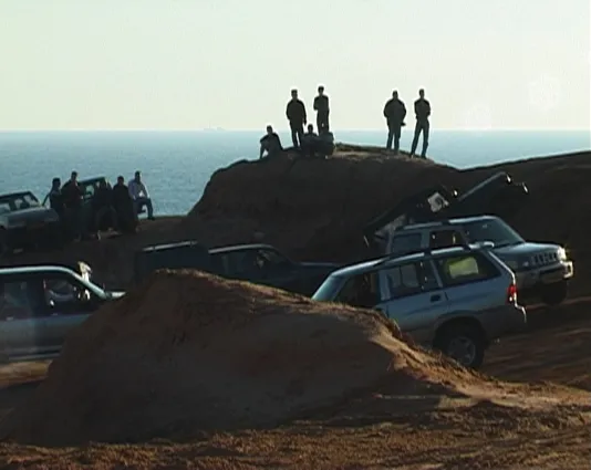 A group of men converge atop coastal dirt hills and nearby their off roading trucks parked around, the ocean is visible behind.