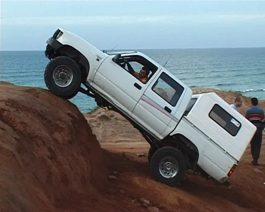 A man attempts to scale a precipitous slope in an off roading truck, the whole front is angled upward against a coastal hill.