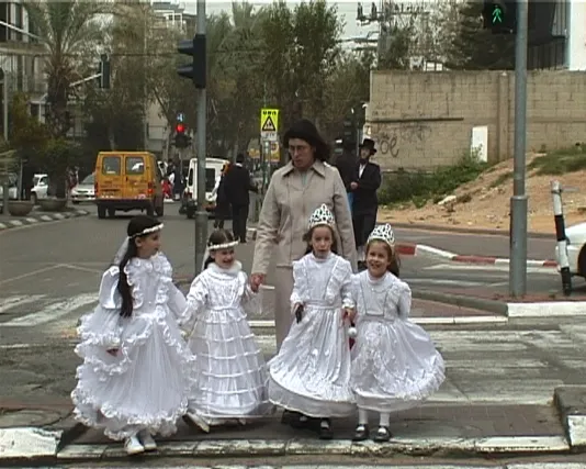 Four merry girls dressed in white gowns, veils and tiaras stand with a woman accompanying them across a busy urban street.