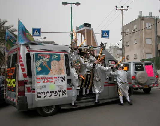 A group of men wearing silver marching band style outfits with shiny hats hang out of a van festooned with flags, signs, speakers.