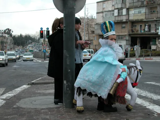A child waits to cross a street dressed in a royal queen costume with a gauzy dress, a foil crown and stuffed riding horse.