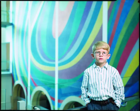 Photo of boy in glasses, a dress shirt, with hands in pockets of pleated pants, posing in front of a colorful abstract mural.
