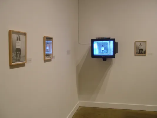 A small monitor hanging alongside wood framed black and white photographs showing the same subject matter on gallery walls.