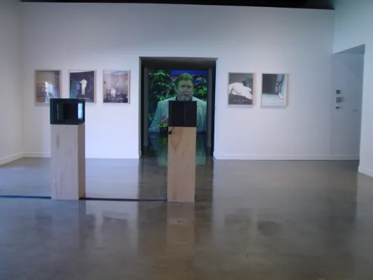 Two video monitors on wood pedestals, a series of photographs flanking a doorway, through which a video projection is visible.