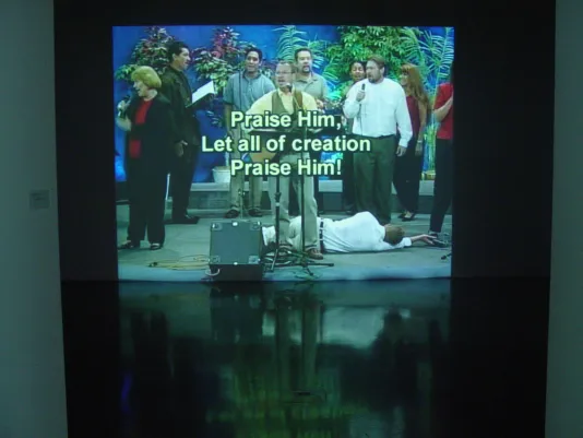 A projected video still of people standing and singing with 1 person lying on a stage, overlaid with a religious message.