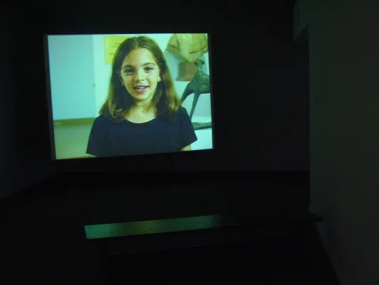 Color video still of a happy young girl speaking, projected in a darkened room with a bench in front.