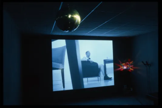 A dark room with a disco mirror ball hung in front of a video projected of a man in a chair with a rainbow disco light beside.