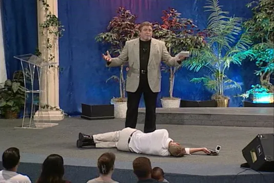 Still of a man lying on his side while another man stands above him with arms open gesturing to an audience from a stage.