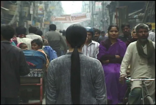 A woman with long black hair dressed in gray clothing stands with her back to the camera on a crowded street in Delhi.