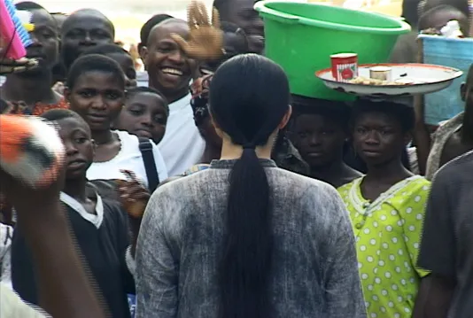 A woman with long black hair dressed in gray clothing stands with her back to the camera on a crowded street in Lagos.