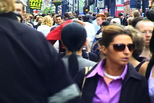 A woman with long black hair dressed in gray clothing stands with her back to the camera on a crowded street in London. 