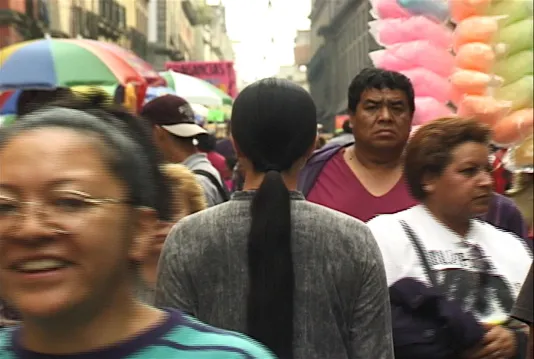 A woman with long black hair dressed in gray clothing stands with her back to the camera on a crowded street in Mexico City.