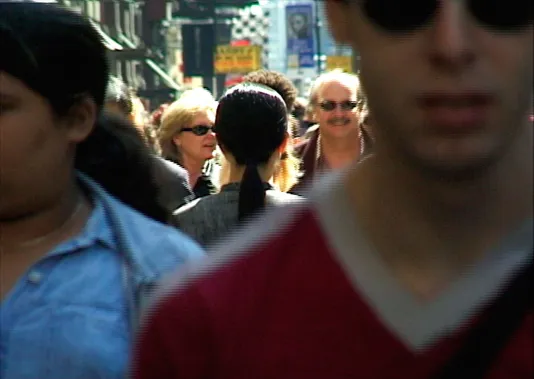 A woman with long black hair dressed in gray clothing stands with her back to the camera on a crowded street in New York City.