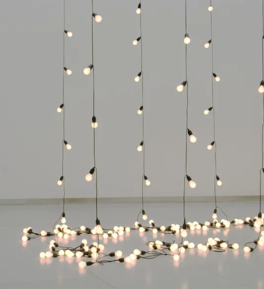 Six strands of small white light bulbs hang down and lie together on the gallery floor.