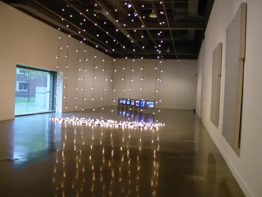Strands of lights hang from ceiling to floor, paintings hang on wall, a line of video monitors on floor, a view thru window.