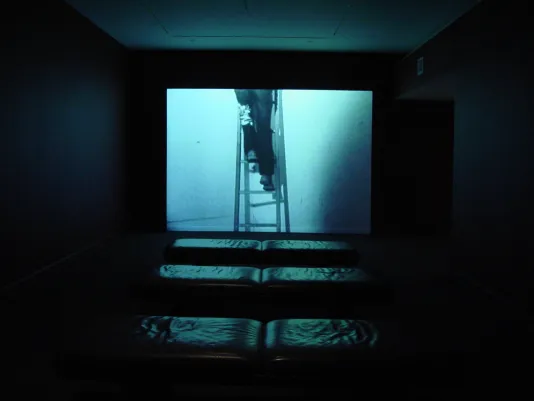 A video of a person's legs standing on a ladder in a blue hue projected in a dark gallery, with a few benches in front.
