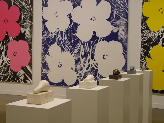A series of lightbulb sculptures lie on pedestals, three large Warhol flower paintings hang on the wall in the background.