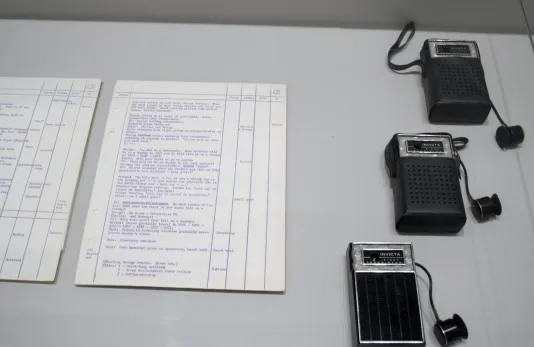 Close view of two handwritten documents and three small vintage hand crank radios arranged under a clear case on a pedestal.