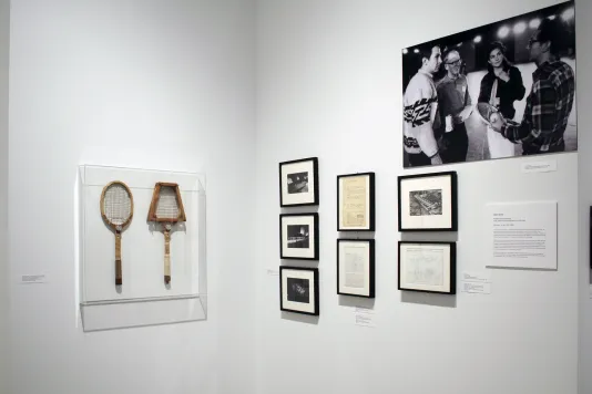 Two tennis rackets in a clear case, a group of small photos, documents, and a large black and white photo hang on two walls.