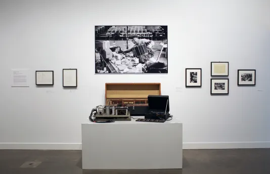 Small black and white photos, documents hang around one large photo above a pedestal displaying vintage electronic objects.