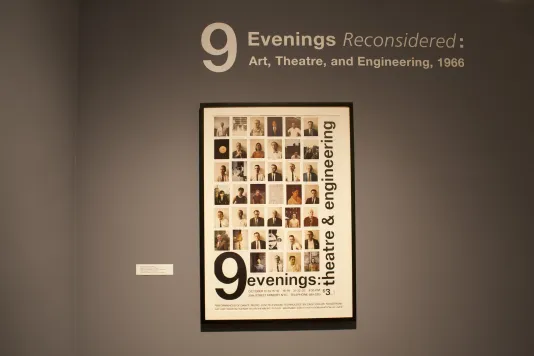 Framed color poster for original 9 Evenings,1966 event hangs on wall below vinyl text that reads 9 Evenings Reconsidered.